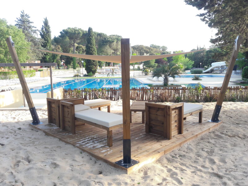 Espace détente avec voile d’ombrage tendue sur poteaux bois, terrasse bois et salon extérieur au bord d’une piscine.